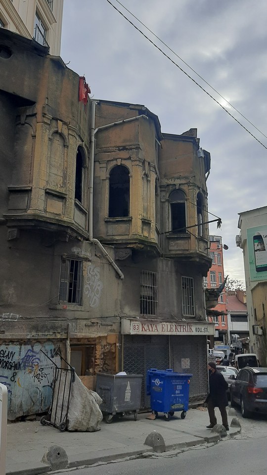 An old, weathered building with arched windows and a shuttered storefront on a narrow street with some trash bins and a pedestrian.