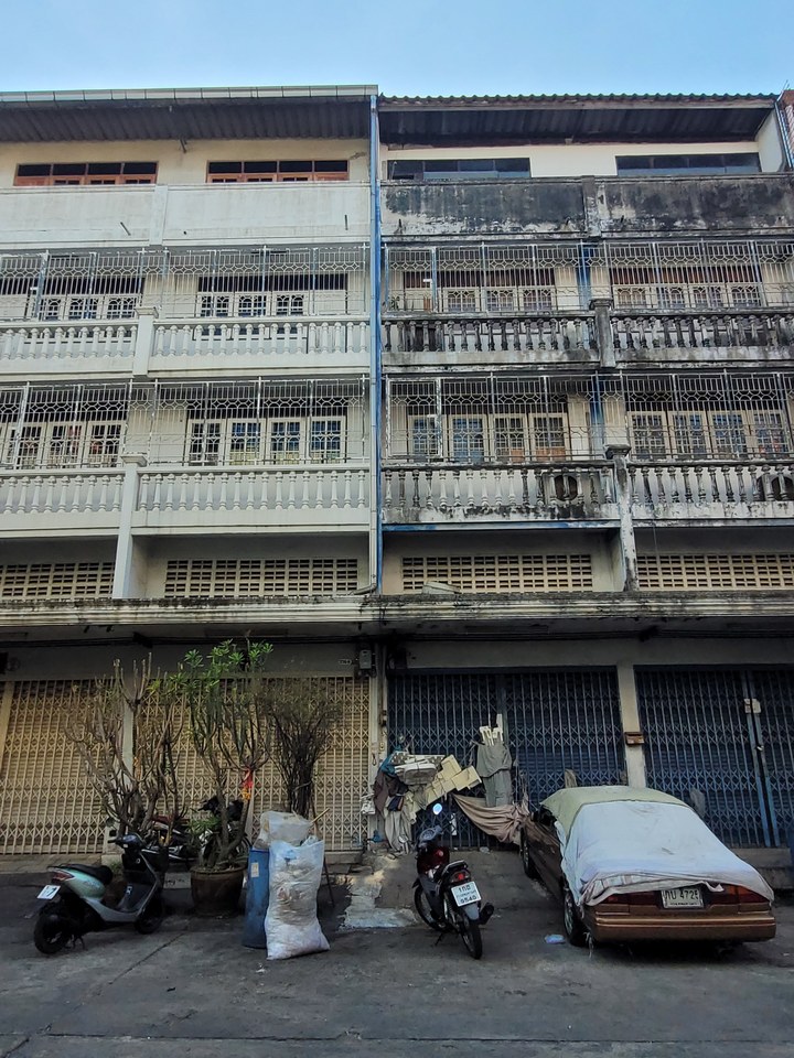 A street view of a worn multi-story building with metal grills, parked scooters, covered car, and potted plants.