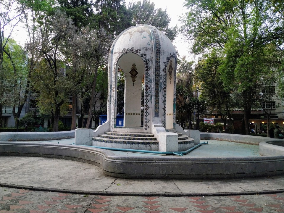 A small, white domed pavilion with decorative tiles stands in the center of a circular concrete basin surrounded by trees and urban buildings.