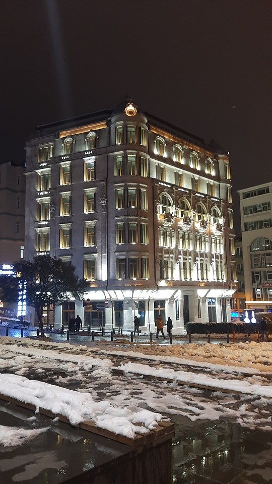 A brightly illuminated historic building at night with snow-covered ground and people walking nearby.