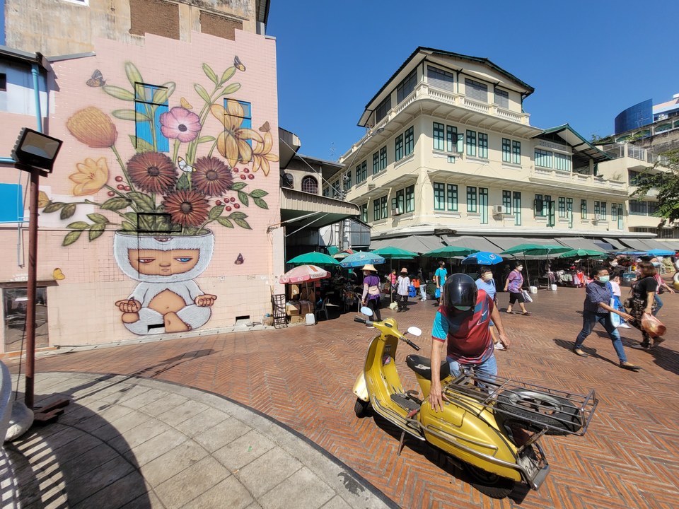 A street scene featuring a large mural of a child with flowers growing from its head, a yellow scooter, and people walking near market stalls.